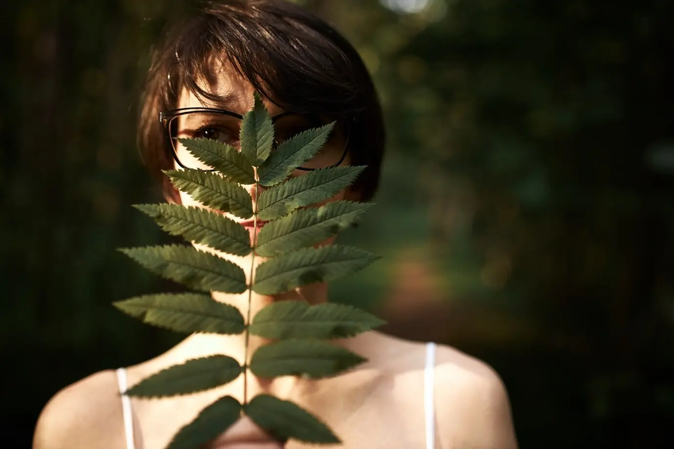 Misteriosa mujer joven de cabello castaño corto y ojos oscuros posando en el bosque, cubriéndose el rostro con una gran hoja verde y disfrutando de la naturaleza salvaje.